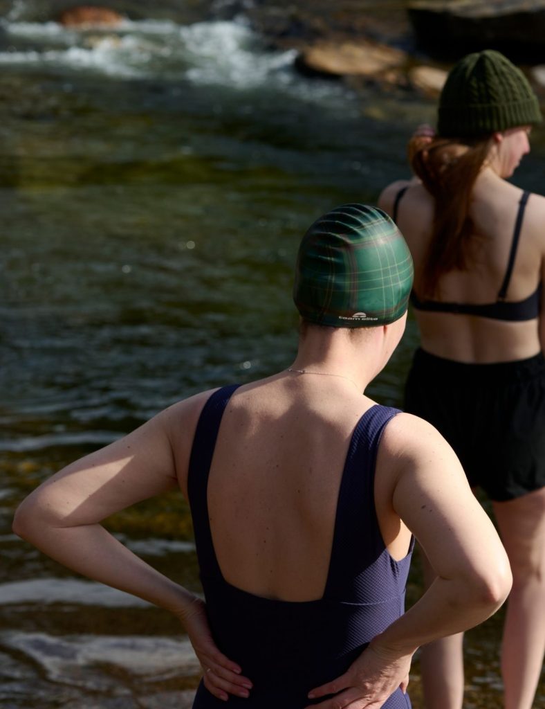 Guest and Annie Armstrong standing at the edge of a river in swim caps and swimwear for a wild swim.