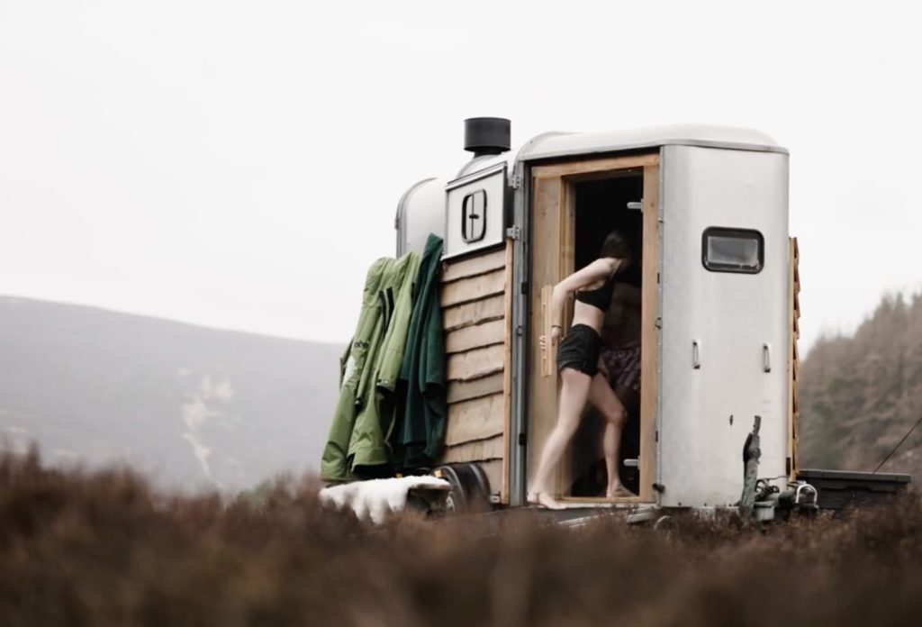 A woman walking into an outdoor sauna housed in a horse box. Coats are hanging on the outside and the Scottish moorland stretches behind.