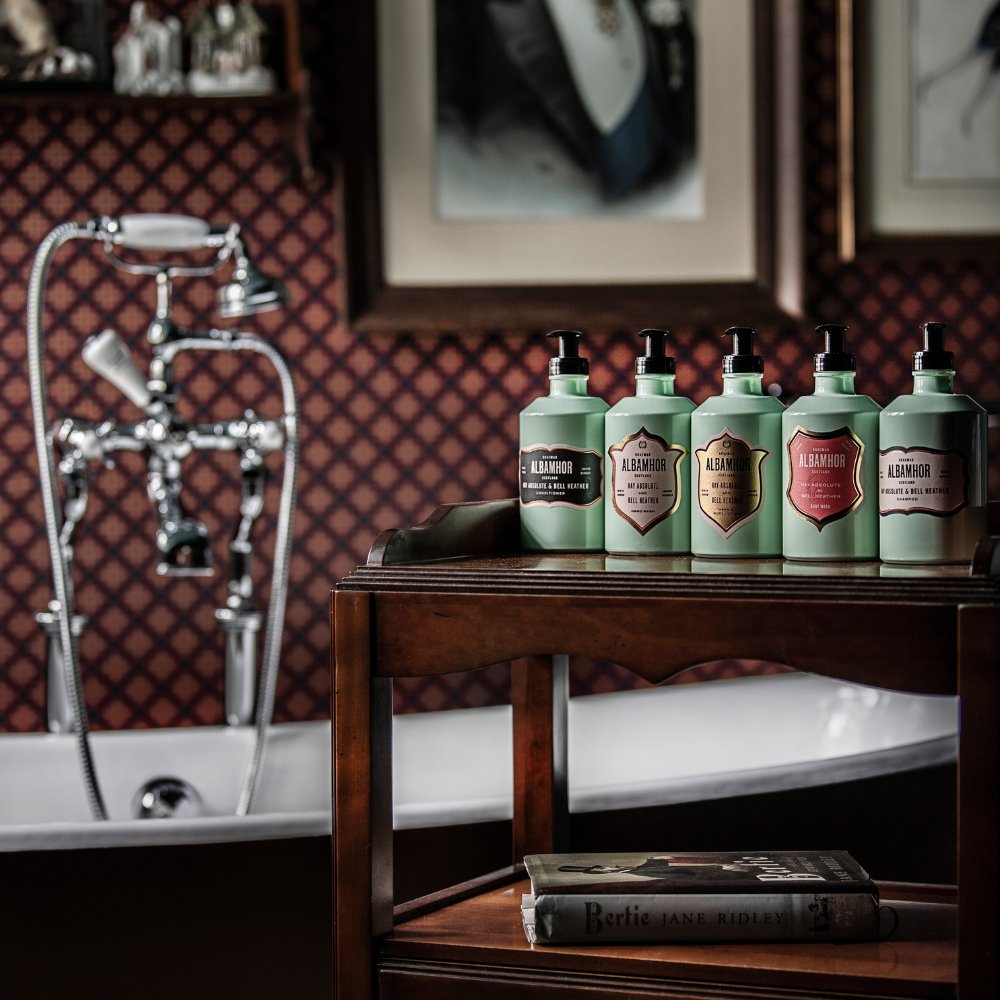 A row of Albamhor pastel‑green toiletries displayed on a wooden stand beside a bathtub, set against patterned wallpaper and framed artwork.