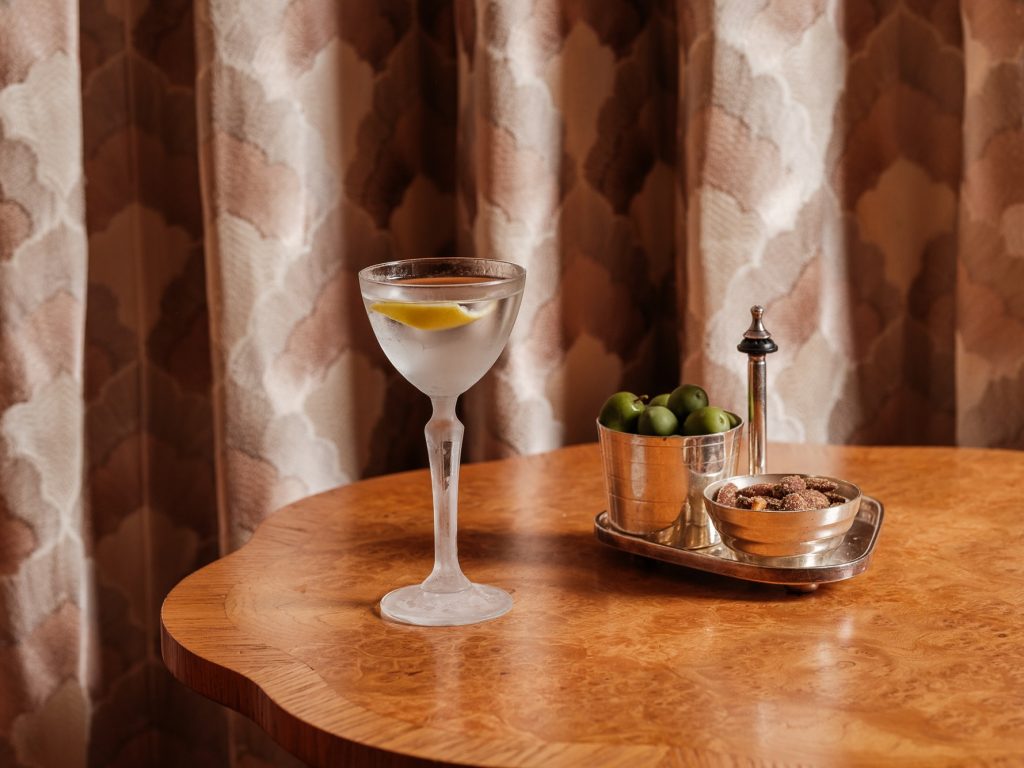 Cocktail glass with a lemon twist beside a tray of olives and nuts on a wooden table in Elsa's Cocktail Bar.