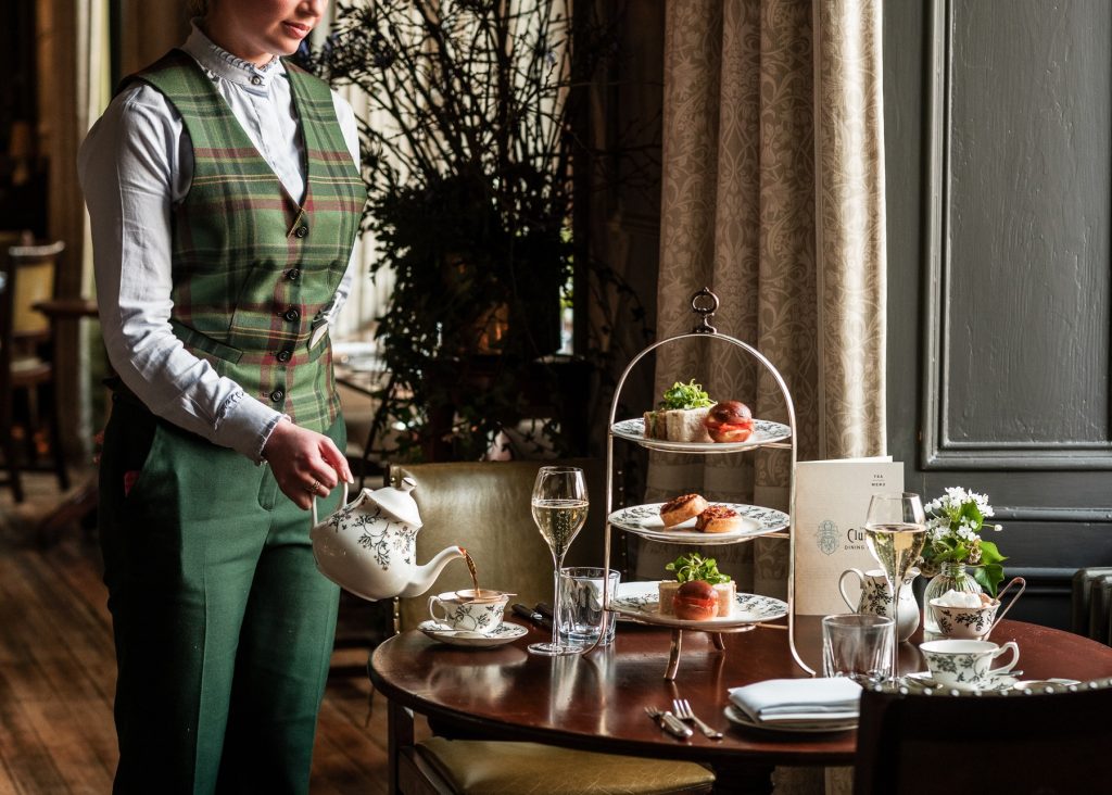 Tea being poured at a table set with a three‑tier stand of sandwiches and pastries in the elegant Clunie Dining Room.