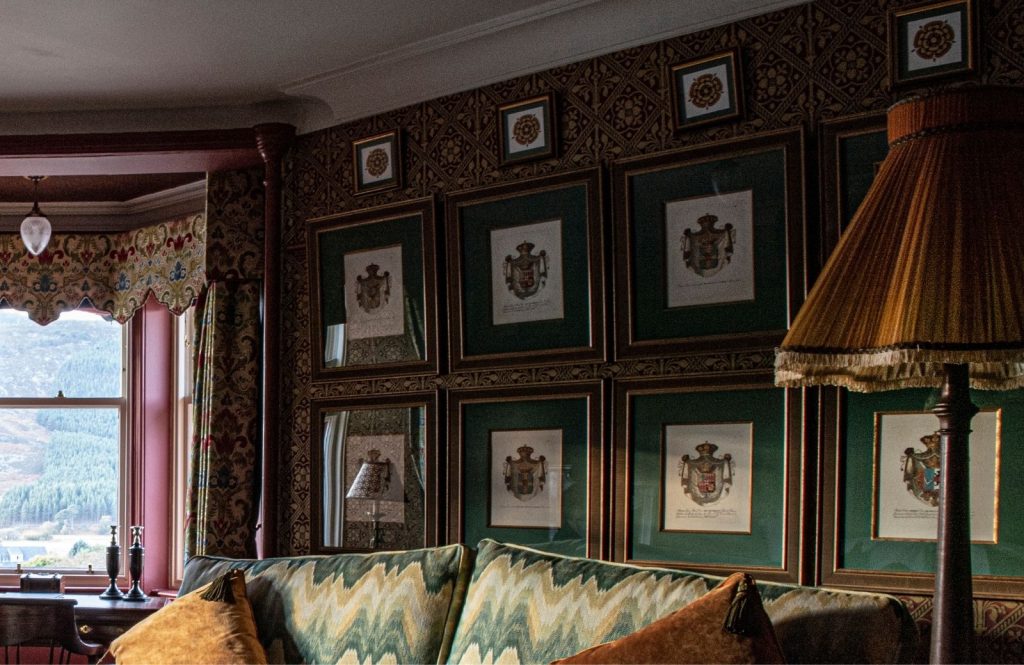 A richly decorated room with patterned wallpaper, framed crests arranged in grids on the wall, a vintage-style lamp, and a window looking out onto a hilly landscape.