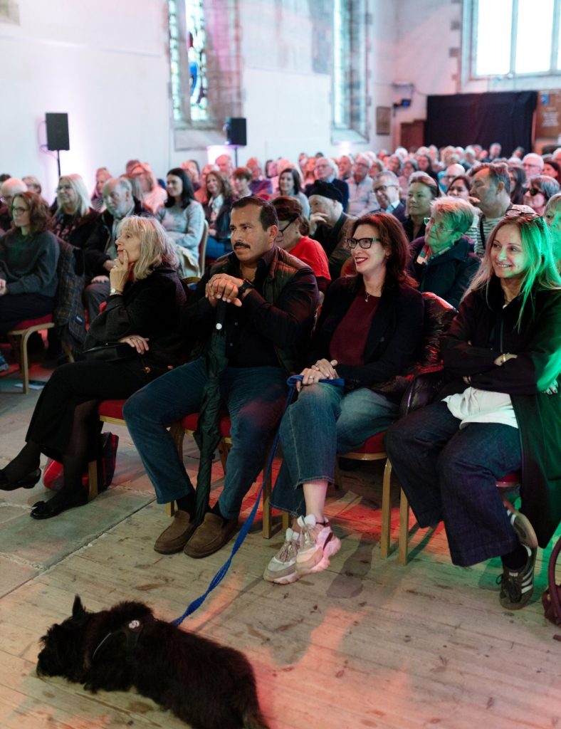 A large audience are sat in rows in a church all looking in the direction of a stage.