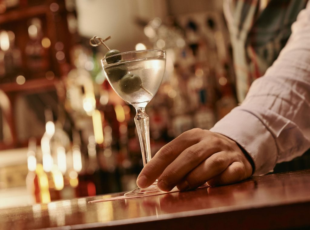 Close-up of a hand placing a martini glass with olives on the bar at Elsa's Cocktail Bar, with blurred bottles in the background.