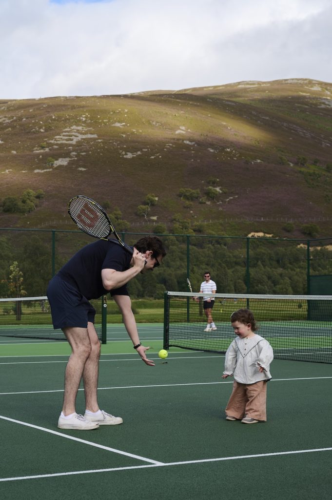 Man throwing a tennis ball to a small child on a tennis court with mountains in the background.