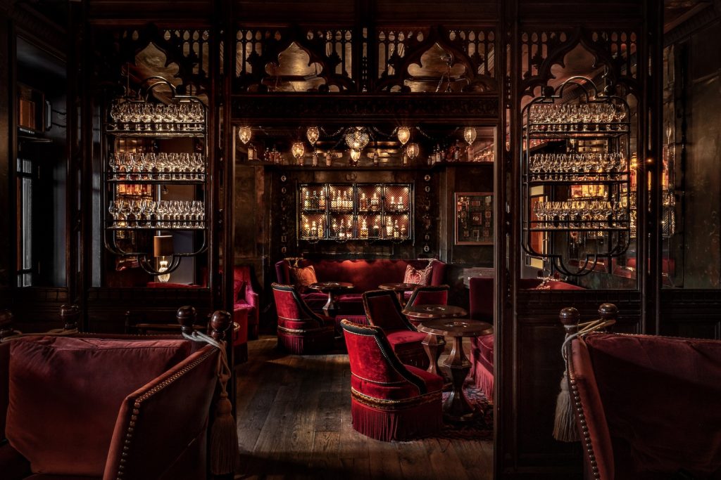 Luxurious dim‑lit Bertie's Whisky Bar with red velvet chairs, ornate dark wood panels, and shelves filled with glassware and bottles.