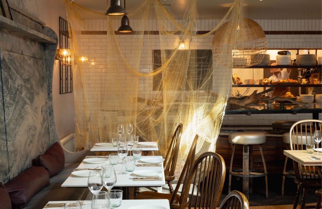 Warmly lit Fish Shop restaurant interior with neatly set tables, wooden chairs, hanging fishing nets, and an open kitchen visible in the background.
