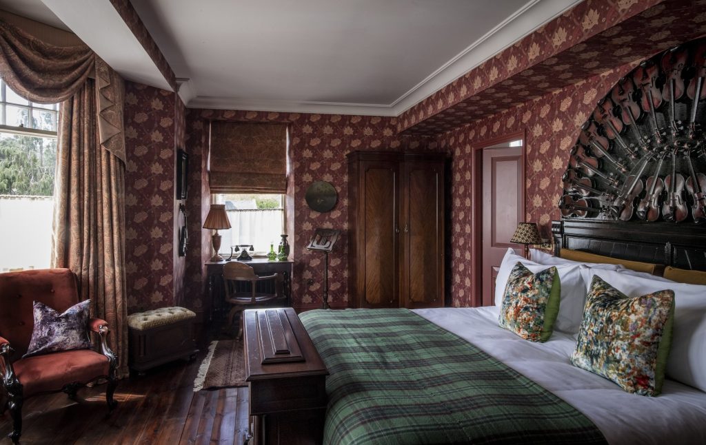 A bed with a tartan throw, red patterned walls, and a headboard made from violins in the Highland Music and Dancing room