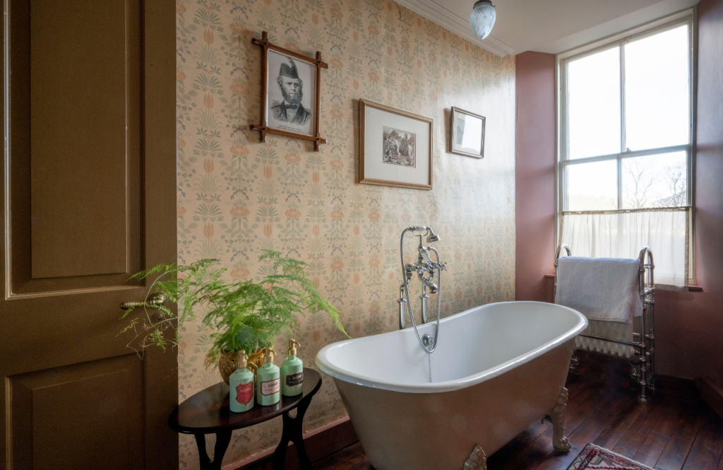 Bathroom in the John Brown suite with a freestanding bathtub, patterned wallpaper, wooden floors and a window with natural light.