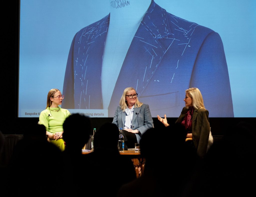 Three female speakers sit on a stage facing an audience with a large screen behind them showing a blue jacket on a manikin covered in white stitches.