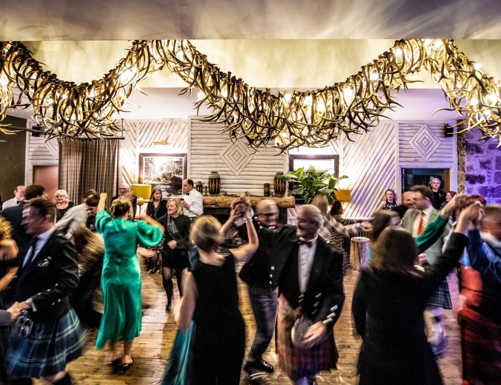 People dancing together in a brightly lit hall decorated with antler-like chandeliers and wooden paneling for Burns Night.