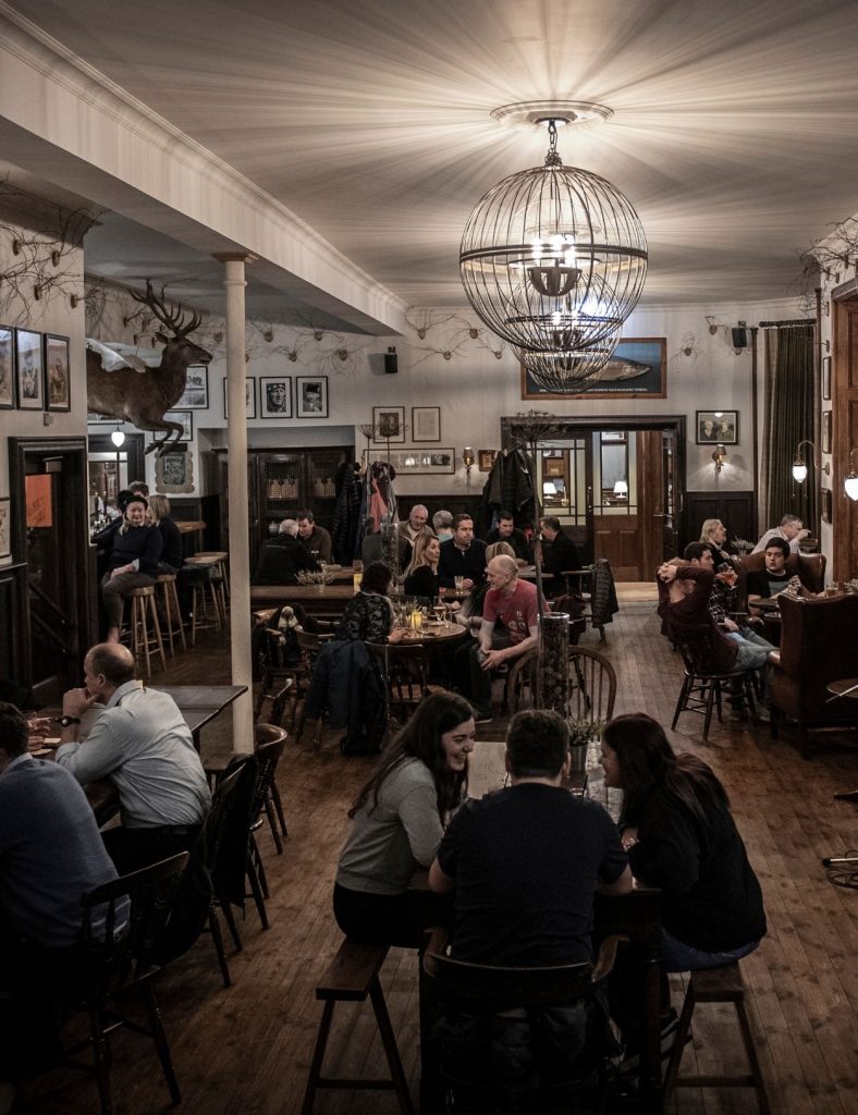 Groups of people seated at wooden tables, talking and drinking under warm lighting and a large spherical chandelier in the Flying Stag pub.
