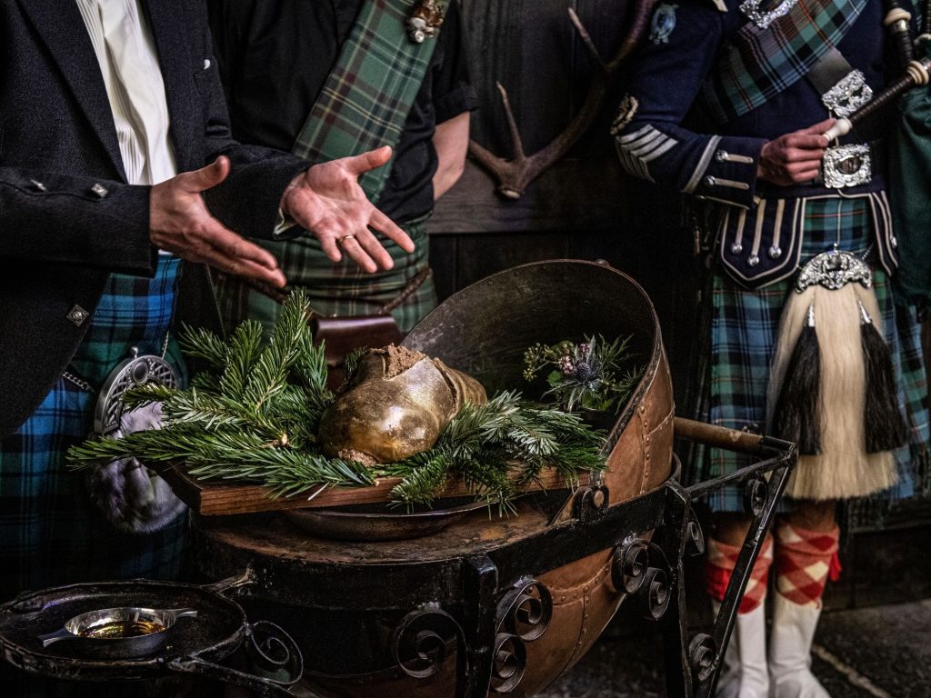 Three people in kilts stand around a haggis sitting on pine needles on Burns Night to address it.