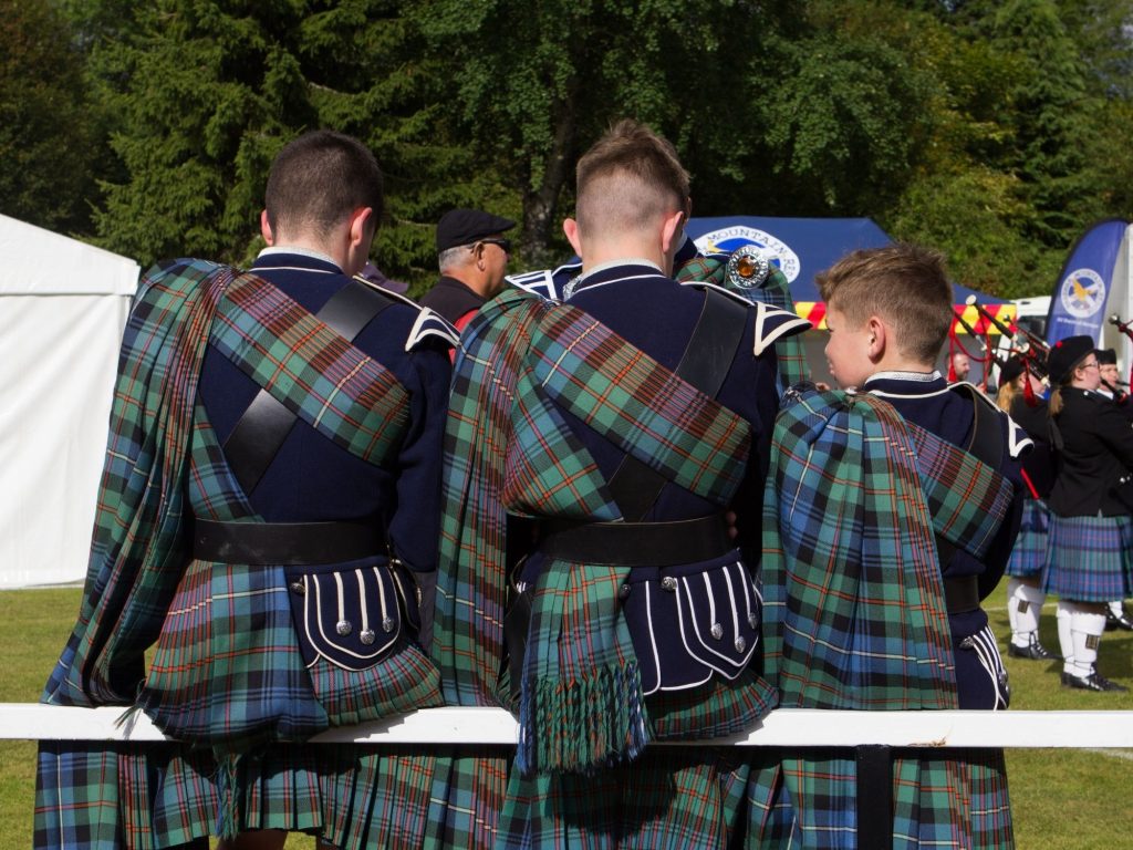 Three Young pipers in matching tartan uniforms leaning on a post with their backs to the camera.