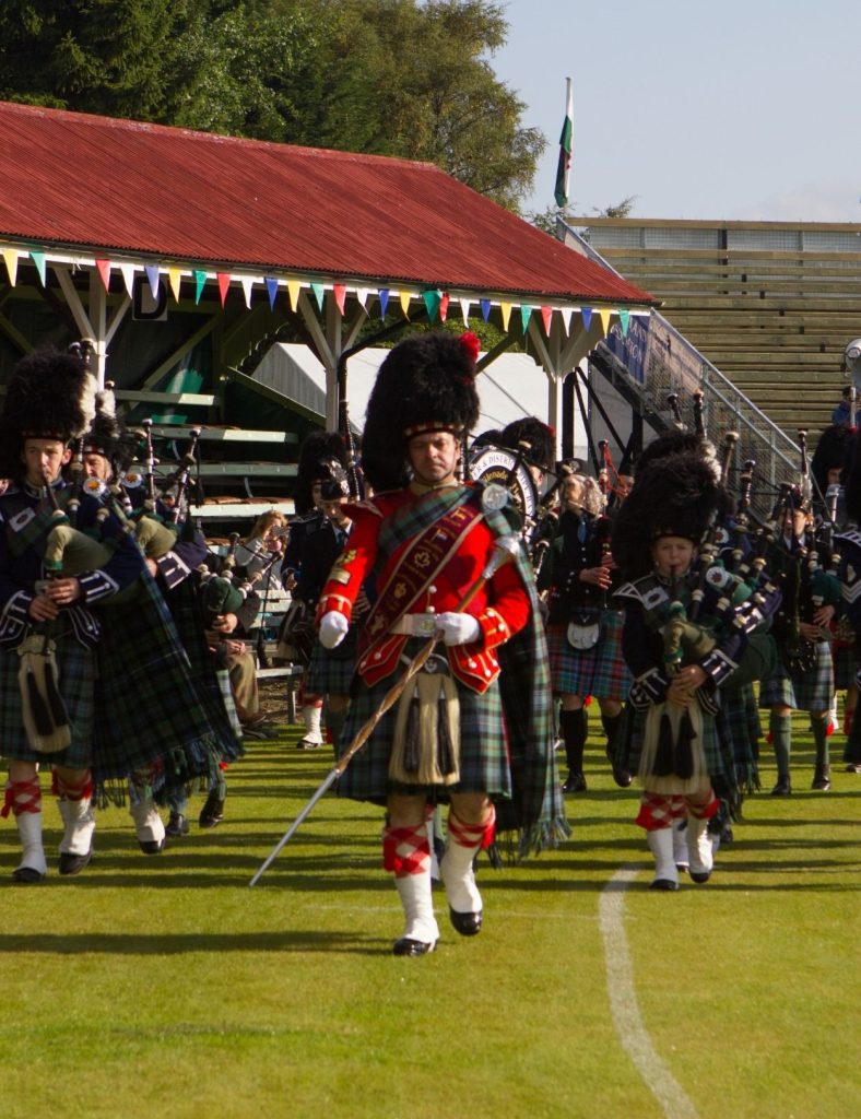 A marching band in traditional Highland dress walking across a grassy field, led by a drum major in a red jacket.