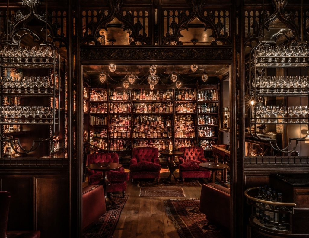 Interior of Bertie's Whisky Bar with dark red velvet arm chairs, dark wood panelling and a wall full of whisky bottles.