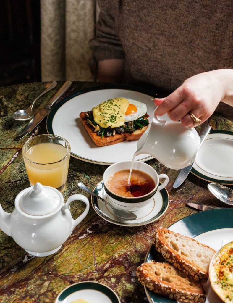 A breakfast table with teapot, milk being poured into a teacup and a plate with toast and an egg on.