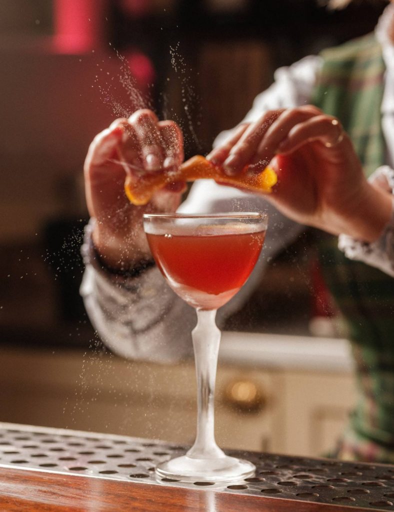 Bartender twisting orange peel over a red drink in a glass, oils from the orange are spraying.