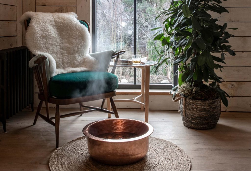 A chair next to a window with a sheepskin throw on the back with a copper bowl of steaming water on the floor.