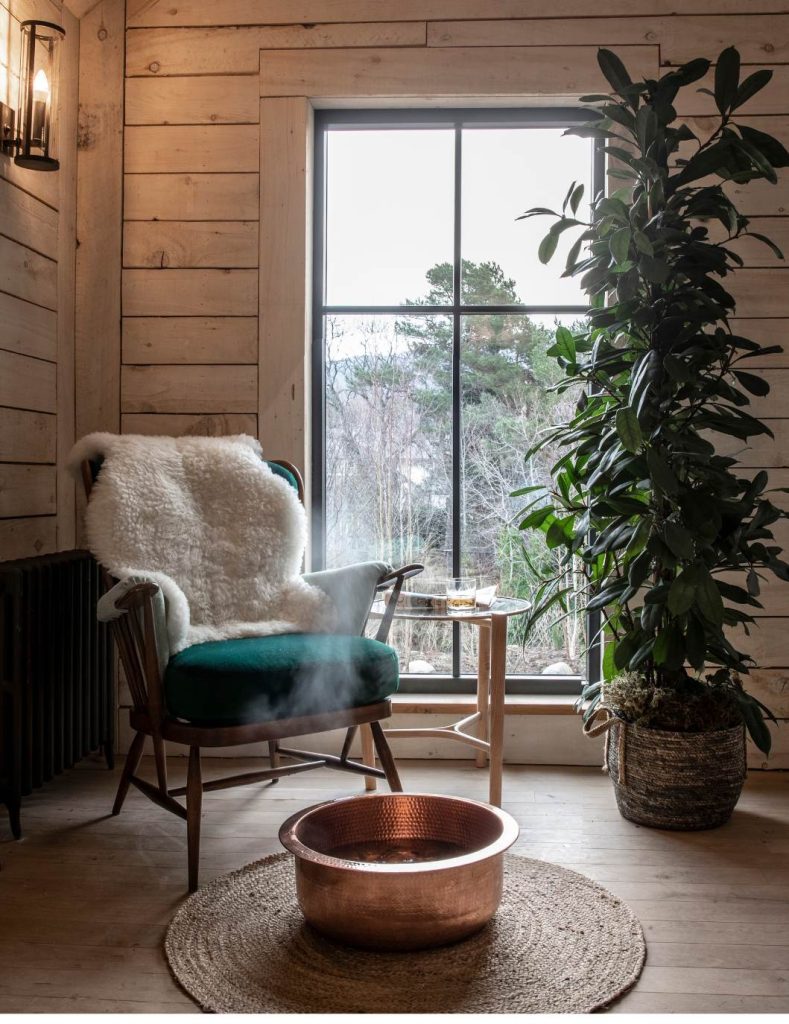 A chair next to a window with a sheepskin throw on the back with a copper bowl of steaming water on the floor.