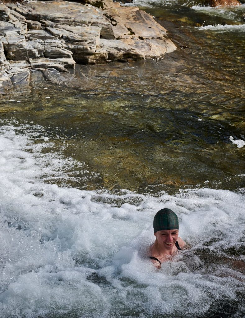 A woman wearing a swimming cap swimming in a river with a waterfall on the left hand side.